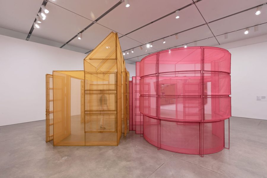 A modern art installation featuring translucent architectural sculptures. One structure is yellow and angular, resembling a house, while the other is pink and curved, resembling a silo or cylindrical tower. Both are set in a white gallery space.