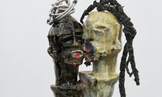 Two abstract, faceless busts—one dark, one yellow—lean together on a pedestal. The dark figure has coiled metal on its head; the yellow figure has twisted black cords as hair. Both have textured, rough surfaces.